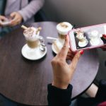 Two women in a cafe capturing photos of their coffee with smartphones. — 8 Advantages Of Using Social Media