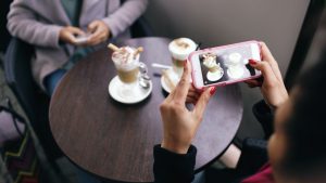 Two women in a cafe capturing photos of their coffee with smartphones. — 8 Advantages Of Using Social Media
