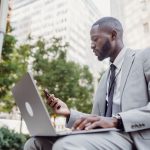 Businessman in a suit using a laptop and smartphone in an urban setting, blending technology with business. — Experience Peac