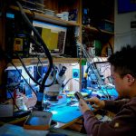 A technician diligently repairs a circuit board at a well-equipped workstation using advanced tools and equipment. — Faceswap