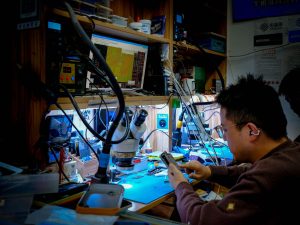 A technician diligently repairs a circuit board at a well-equipped workstation using advanced tools and equipment. — Faceswap