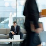 Businessman sitting in modern office lounge using a laptop while colleague walks past, creating dynamic blur. — Powerful Vapo