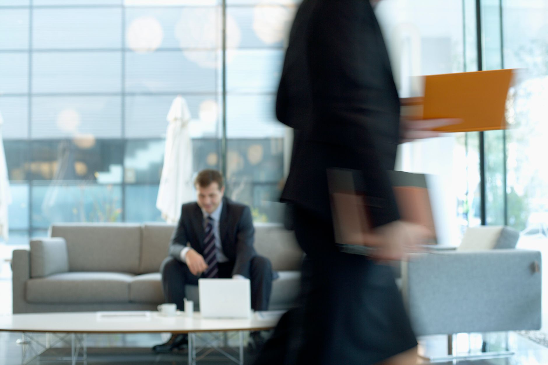 Businessman sitting in modern office lounge using a laptop while colleague walks past, creating dynamic blur. — Powerful Vapo