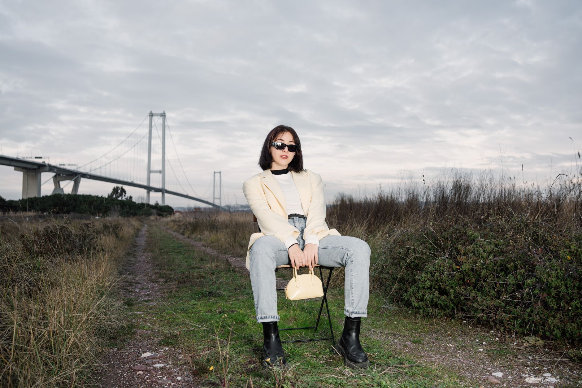Stylish woman in sunglasses sitting by a bridge, showcasing modern fashion against a natural backdrop. — Analyzing Op