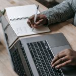 Crop unrecognizable African American female in formal wear writing notes in notepad while working on laptop — Unlock the Powe